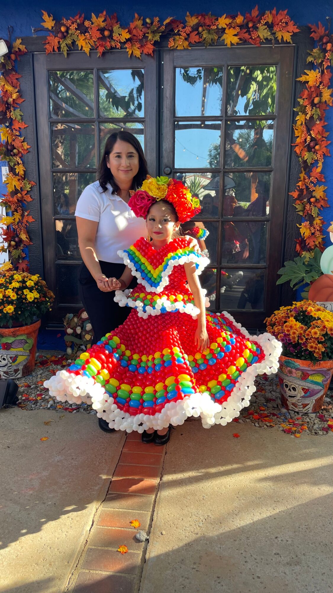 Two people, one in a colorful dress, stand in front of a window with autumn decorations and potted flowers.