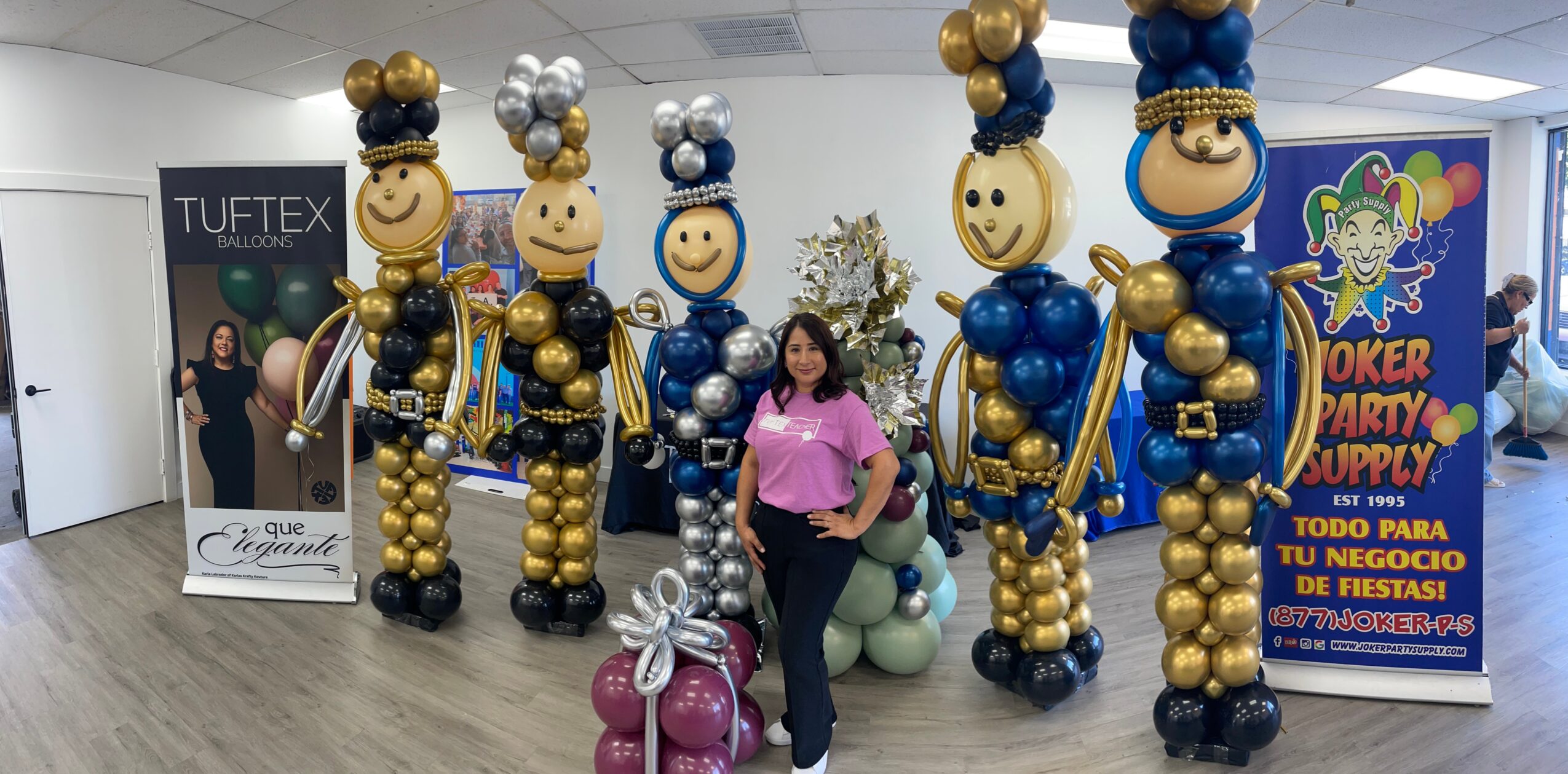 Woman standing among large balloon figures dressed as clowns, with promotional banners in the background.