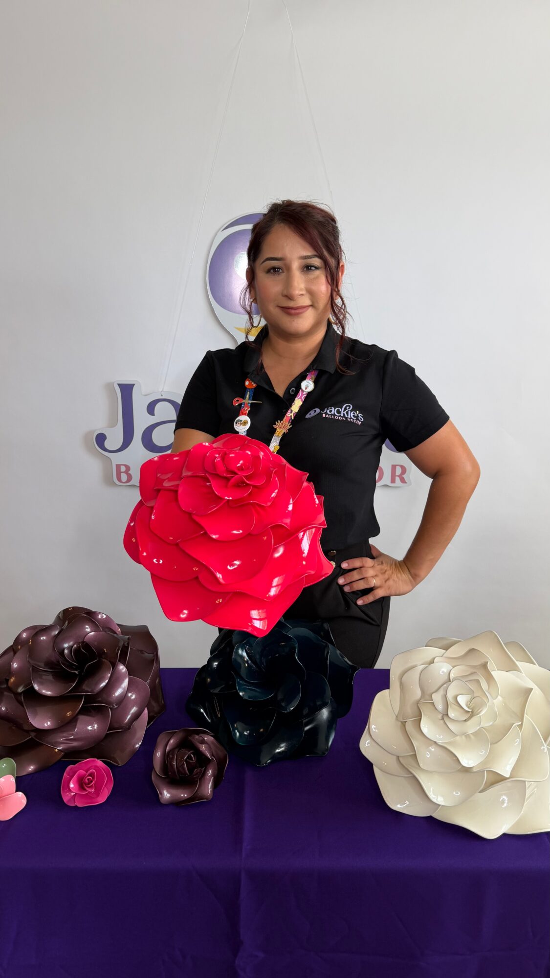 Woman standing behind table with large paper flowers, holding a red flower, smiling, white wall background.