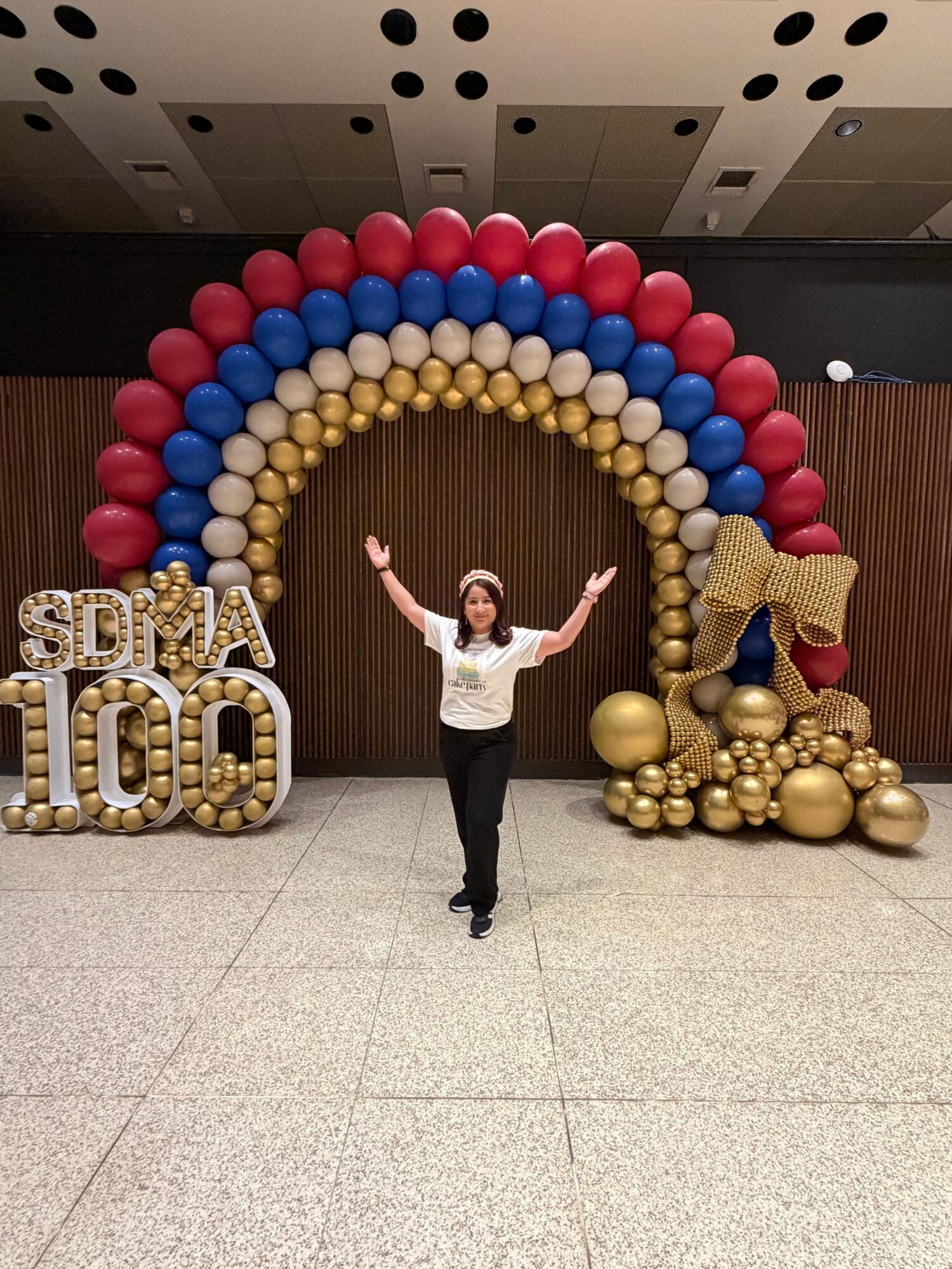 Person standing in front of a balloon arch with gold, red, and blue balloons, and a large gold '2023' sign.