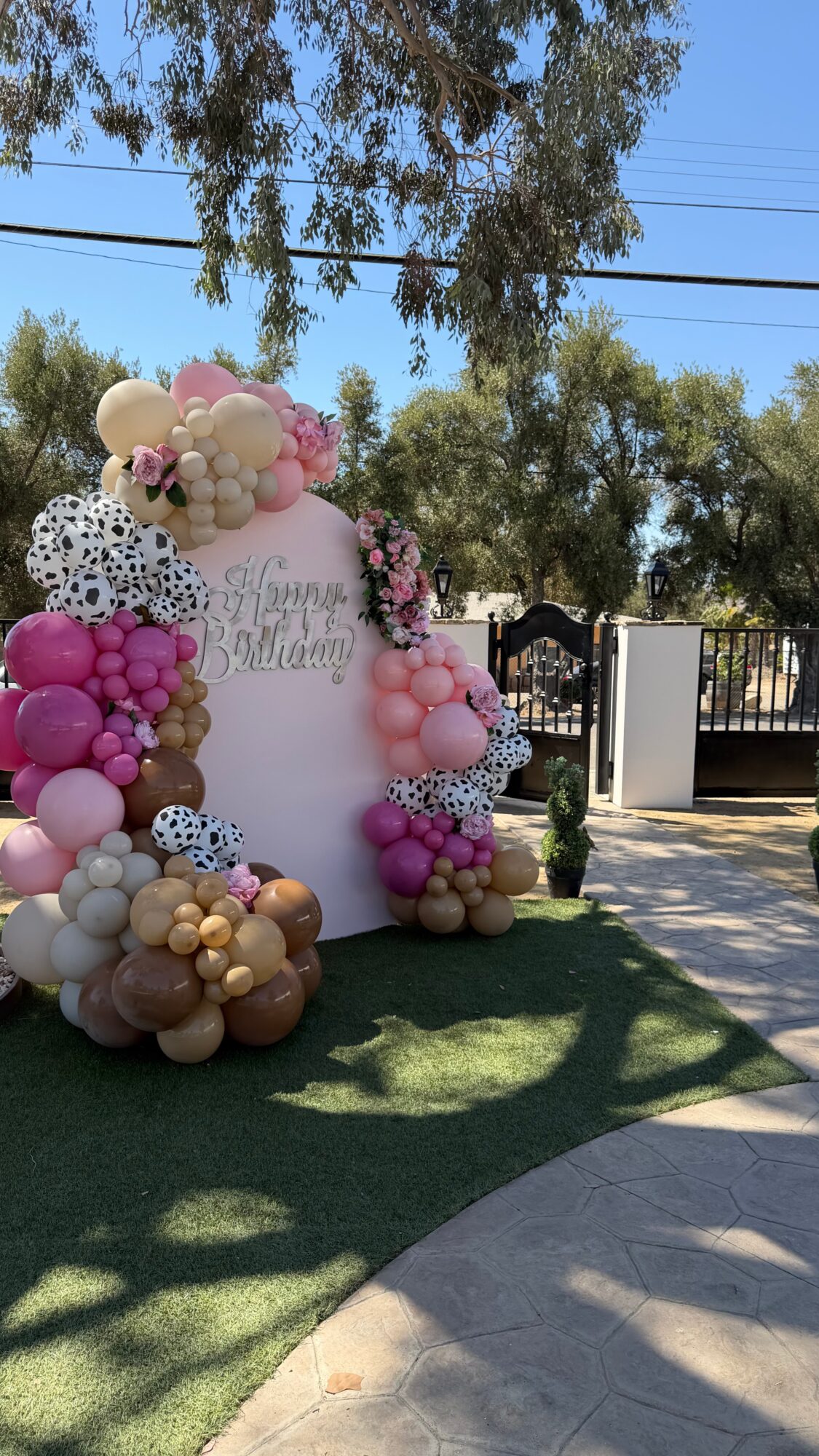 Balloon arch with pink, white, black, and gold balloons, and a sign reading 'Happy Baby Shower' in a yard with trees and a fence