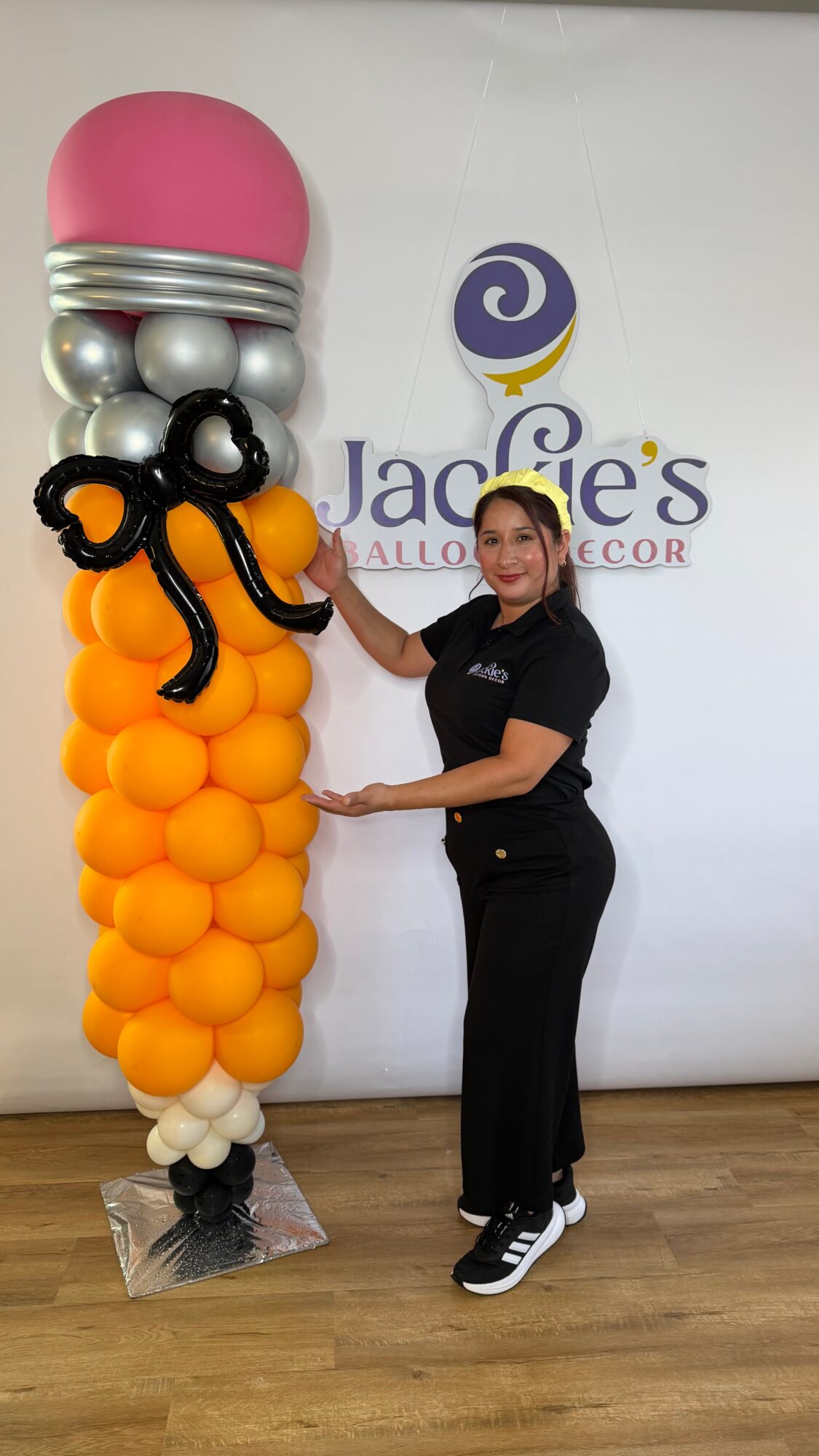 Woman standing next to a tall balloon sculpture with pink, silver, black, orange, and white balloons, in front of a white wall with a logo.