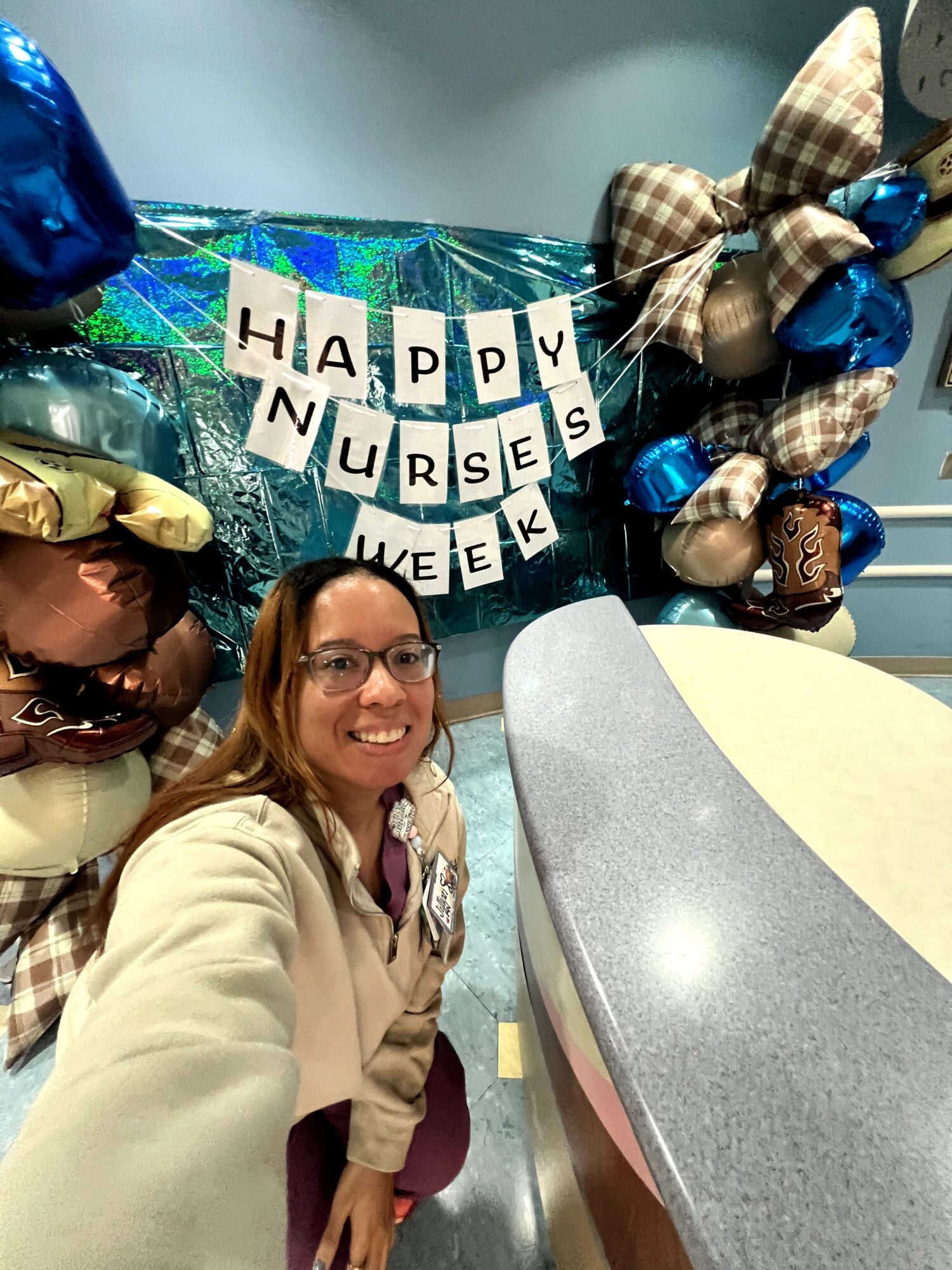 Smiling woman in beige coat taking selfie in front of nurse appreciation decorations with balloons and a banner reading 'Happy Nurse Week'.