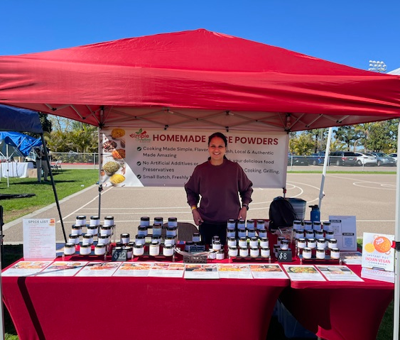 Woman standing behind a table with jars and products under a red canopy at an outdoor market.