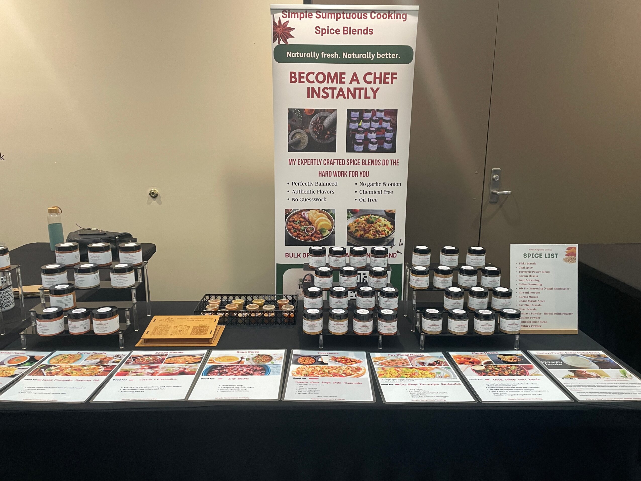 Display table with spice jars, promotional banner, and printed recipes or flyers, set against a plain wall.