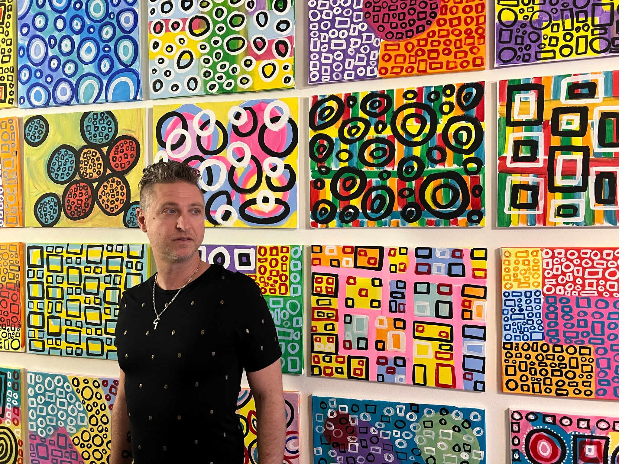 Man in black shirt stands in front of colorful abstract artwork with geometric patterns.