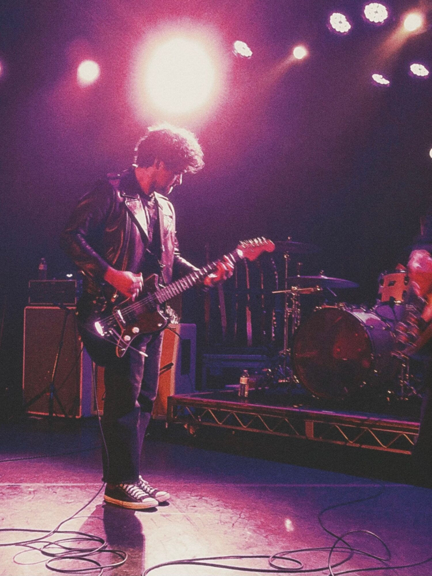 Person playing electric guitar on stage with bright lights overhead, wearing a leather jacket and sneakers.