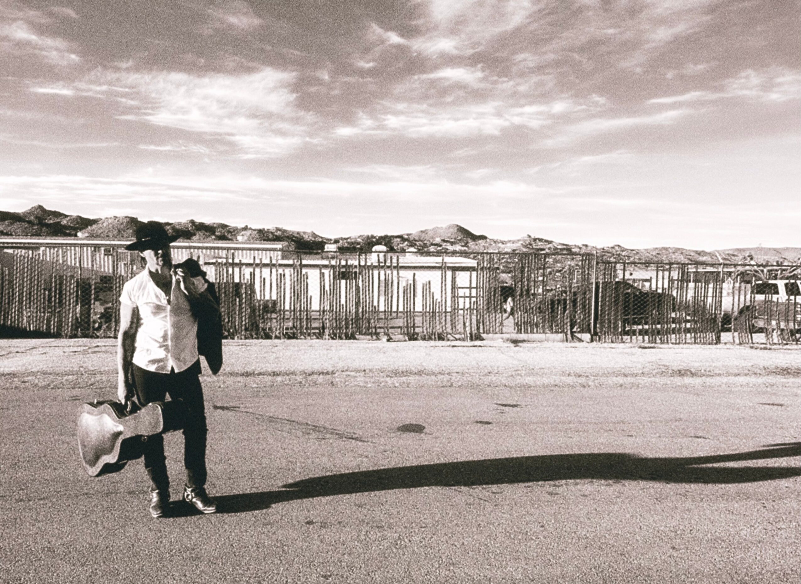 Person walking on street carrying a guitar case, with a fence and hills in background, under cloudy sky