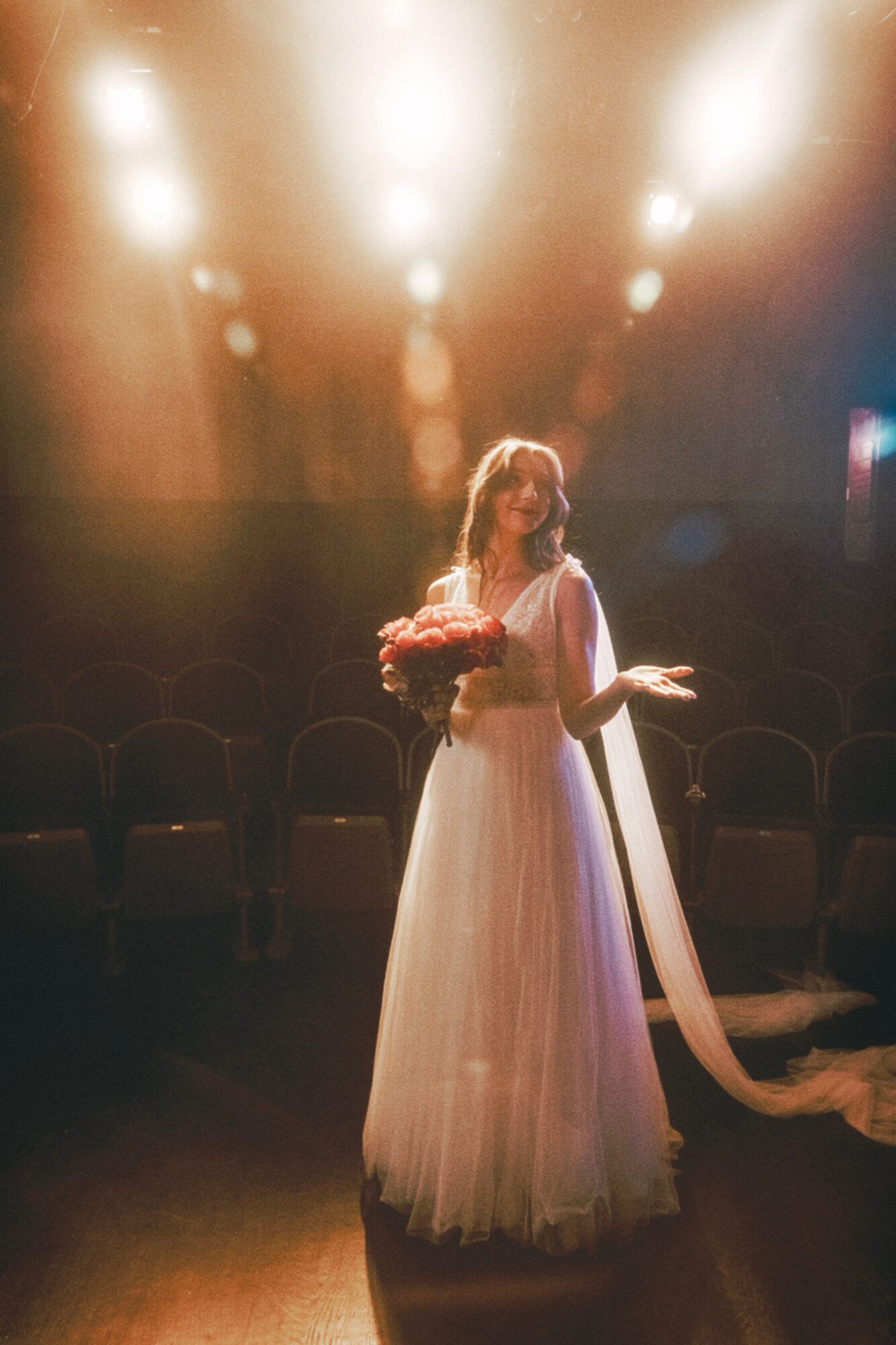 Woman in a long dress holding a bouquet, standing on a stage with bright lights overhead.