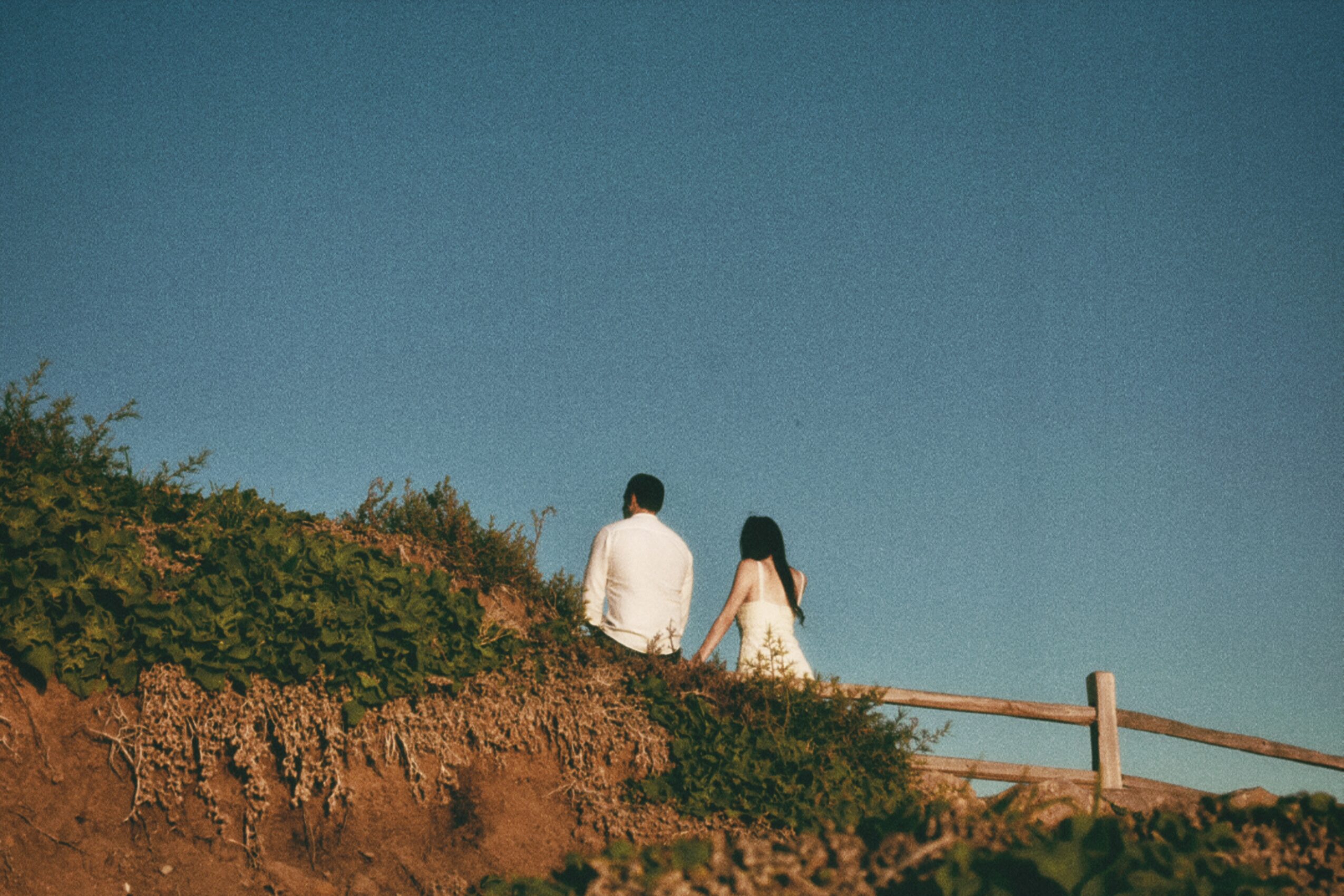 Two people walk along a path with a wooden fence, surrounded by greenery and a clear blue sky.