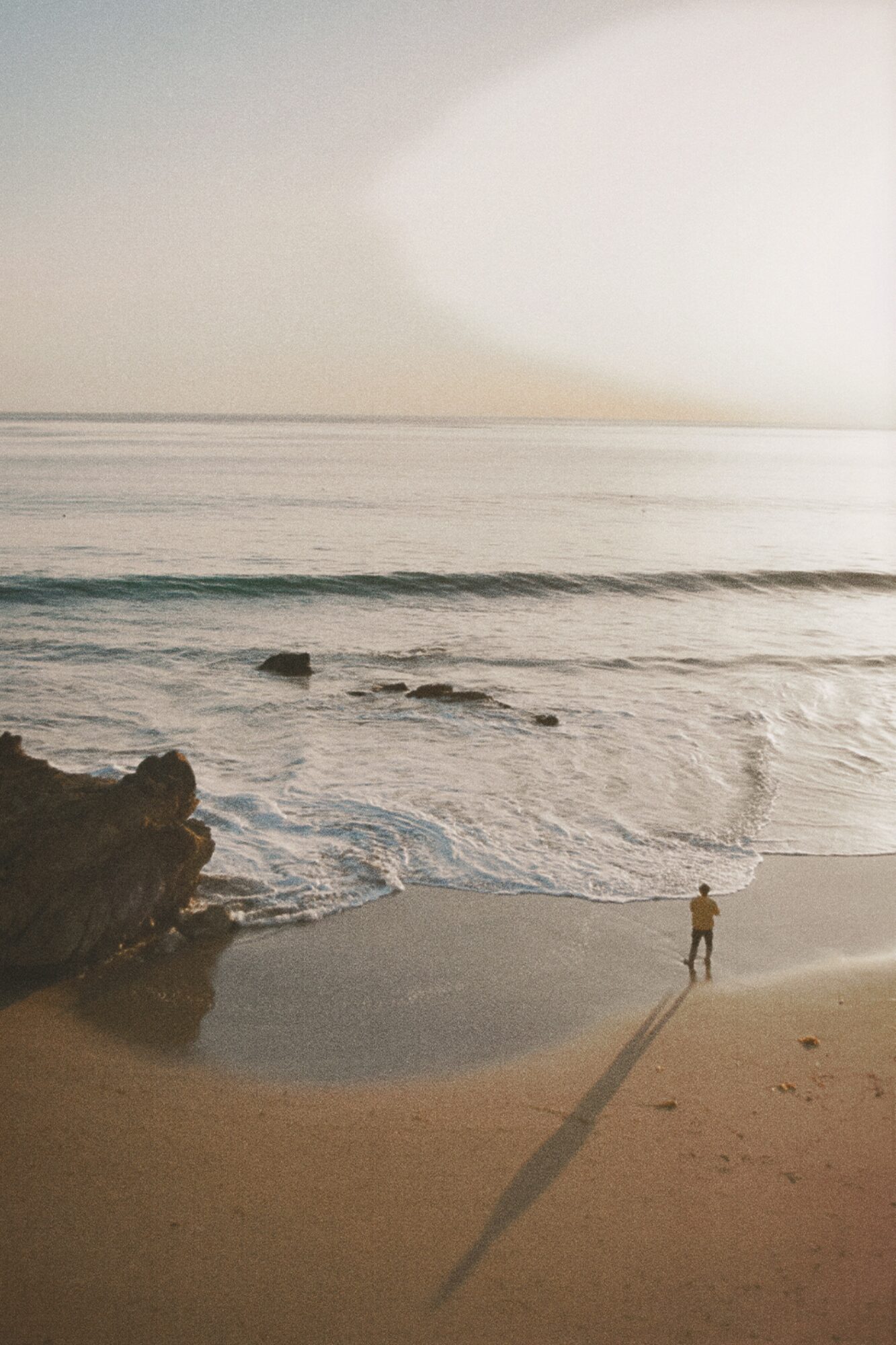 Person standing on beach near water with a long shadow, rocks on the left, sky above, and calm ocean waves.