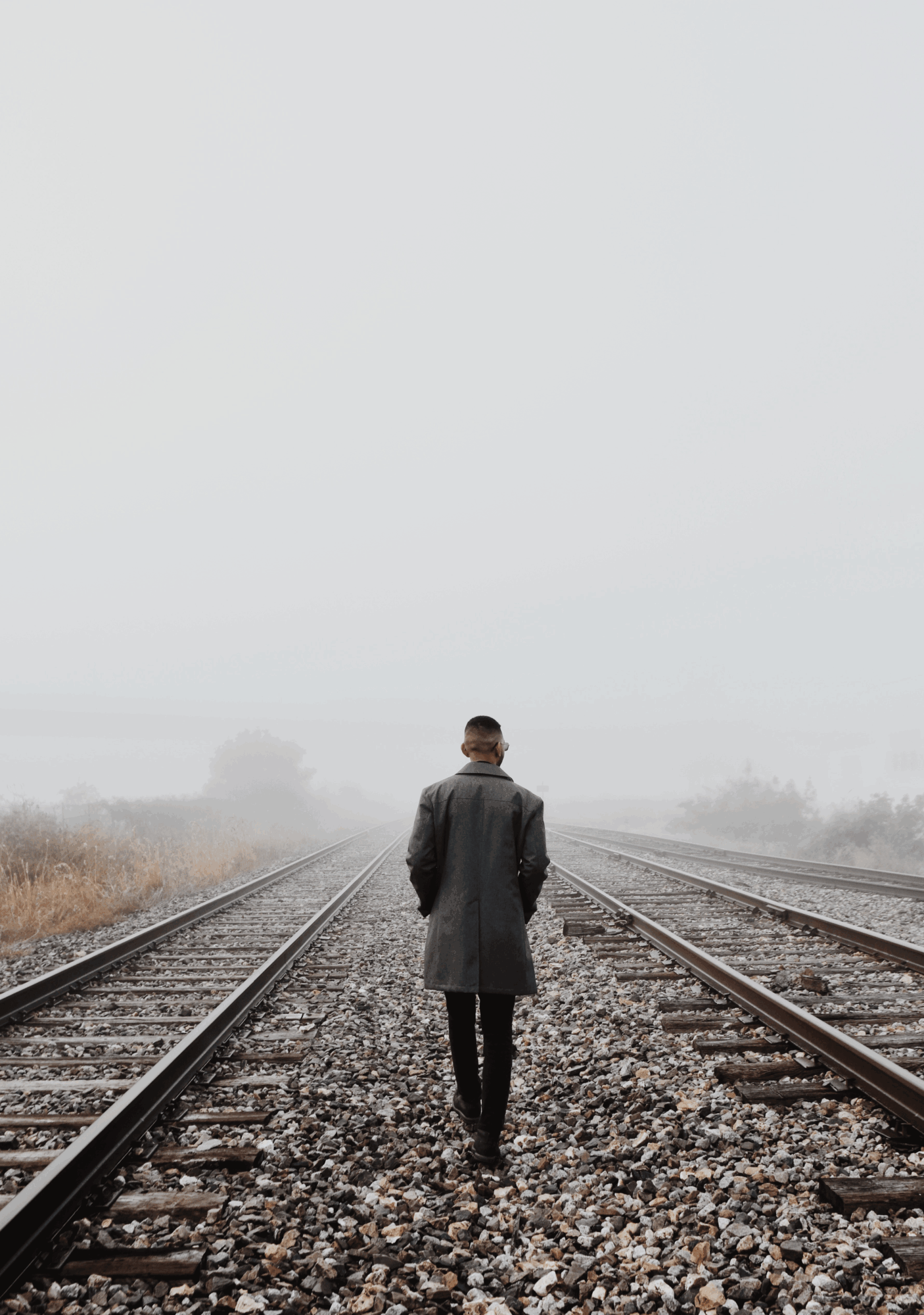 Person walking along train tracks in foggy landscape, wearing a coat and facing away from the camera.