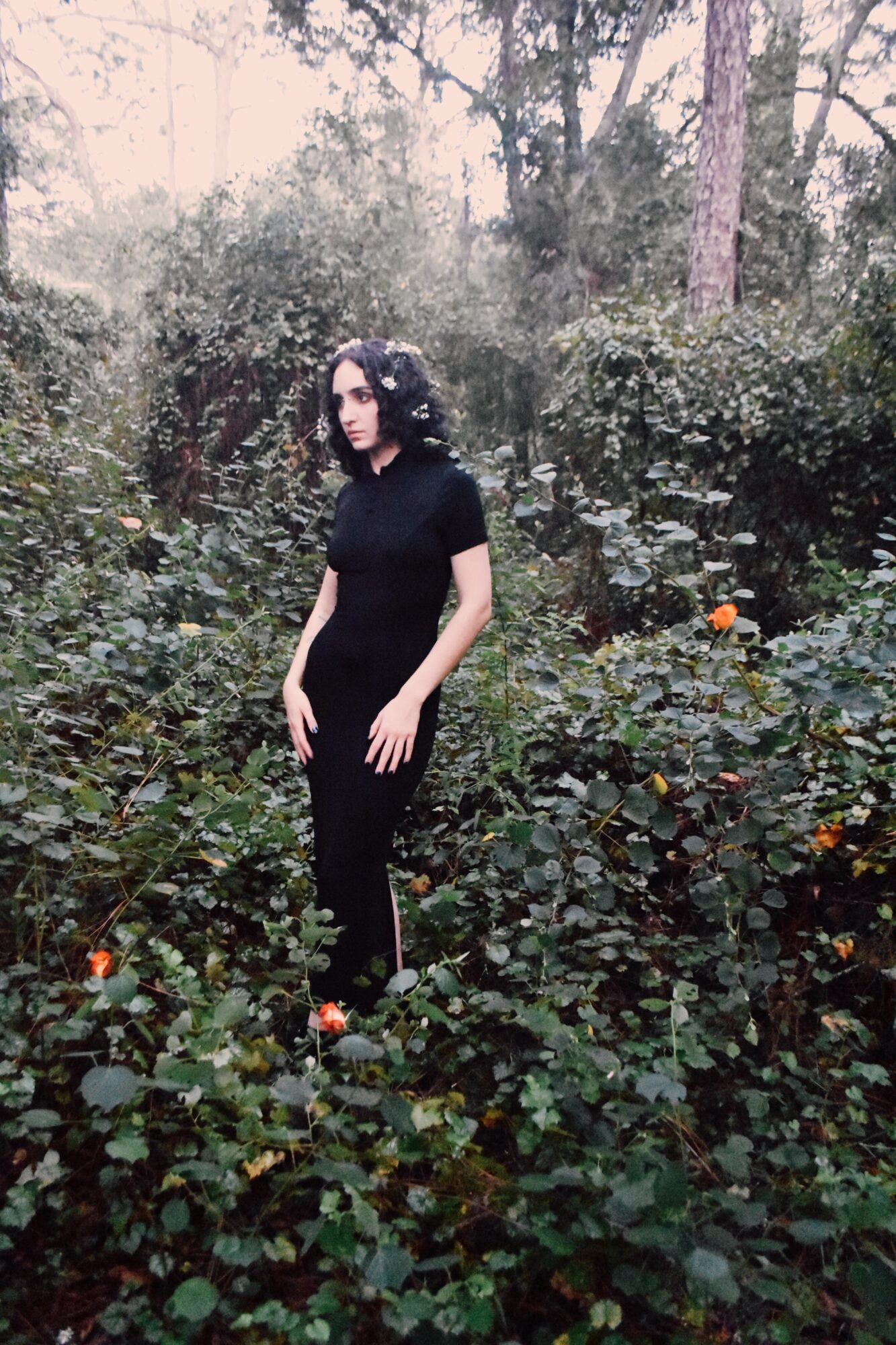 Woman with dark curly hair in black clothing standing in a lush garden with green foliage and small orange flowers.
