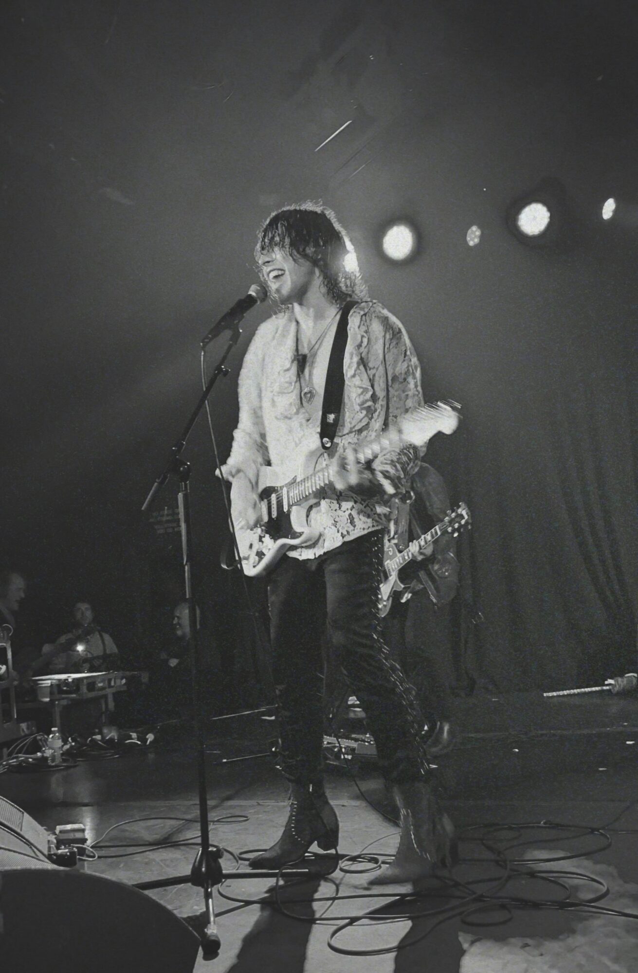Musician singing into microphone while playing electric guitar on stage with bright lights behind.