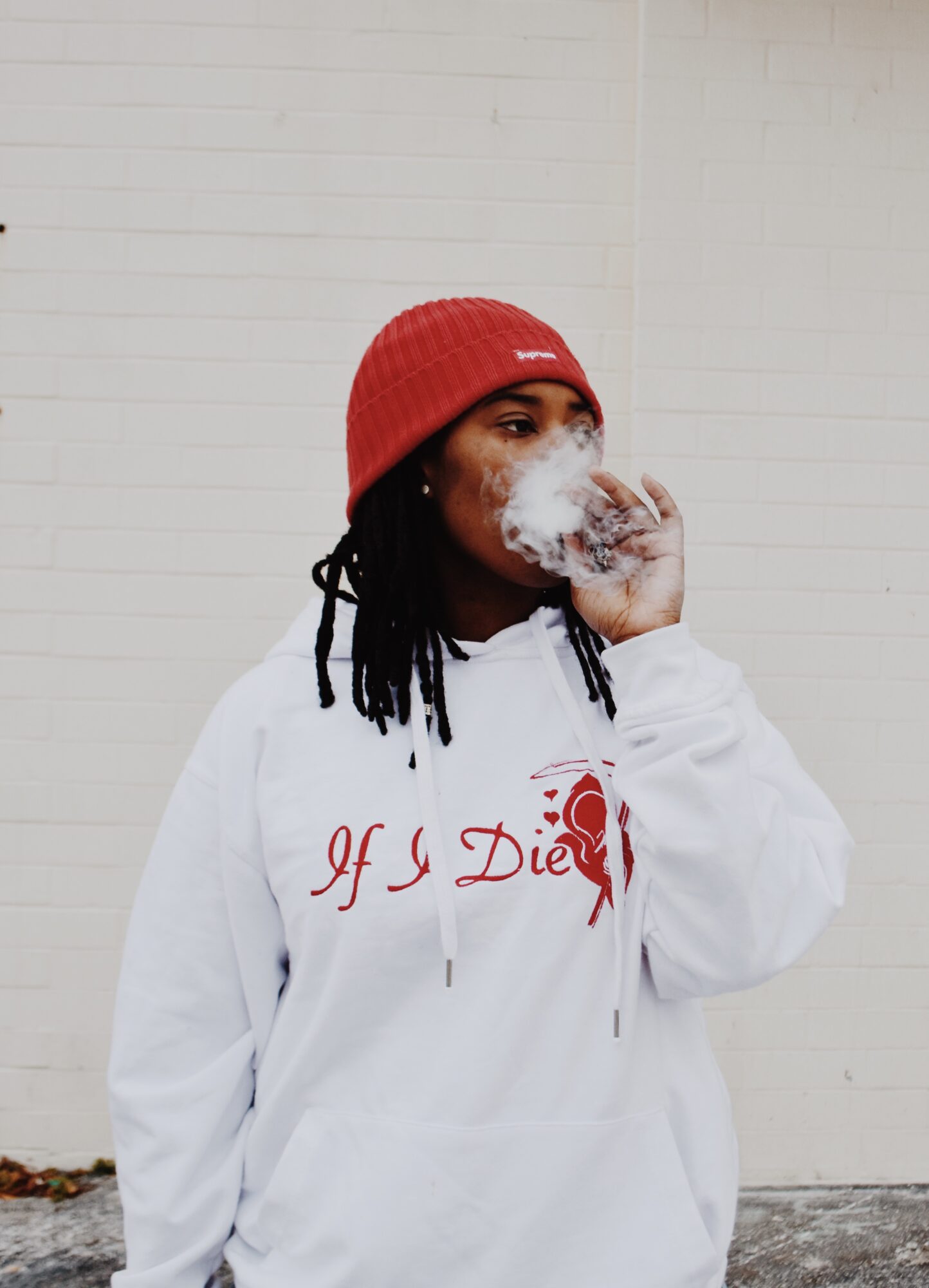Person with dreadlocks wearing a red beanie and white hoodie, smoking, standing against a plain wall.