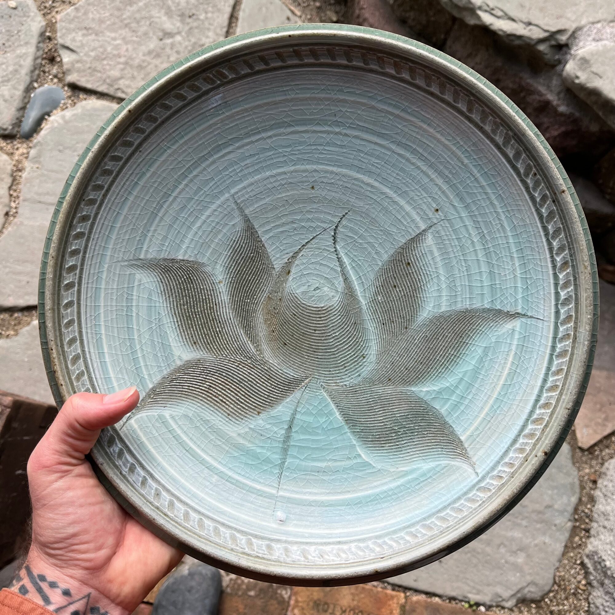 Hand holding a round ceramic plate with a blue and gray floral pattern, outdoors on stone surface.