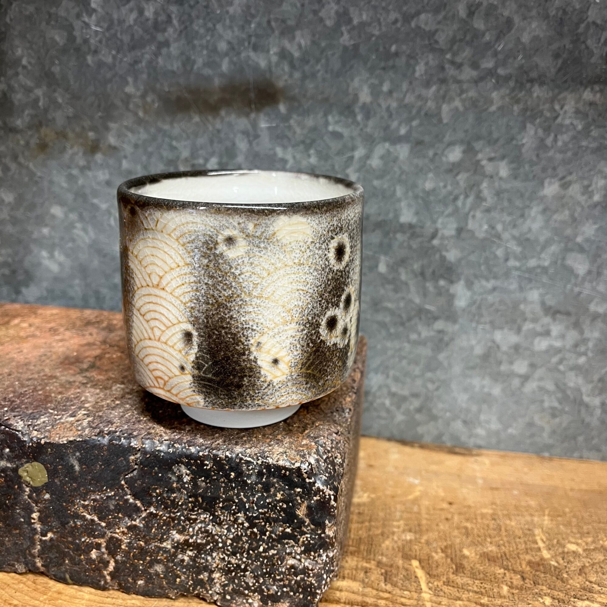 Small ceramic cup with black and white patterns, placed on a rough stone surface against a gray background.