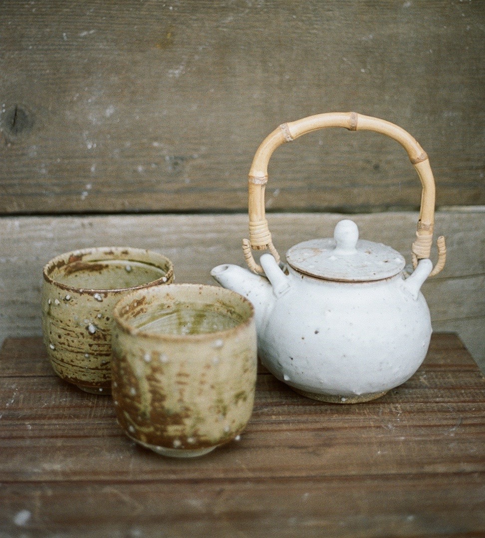 White teapot with a bamboo handle and two ceramic cups on a wooden surface, against a wooden background.