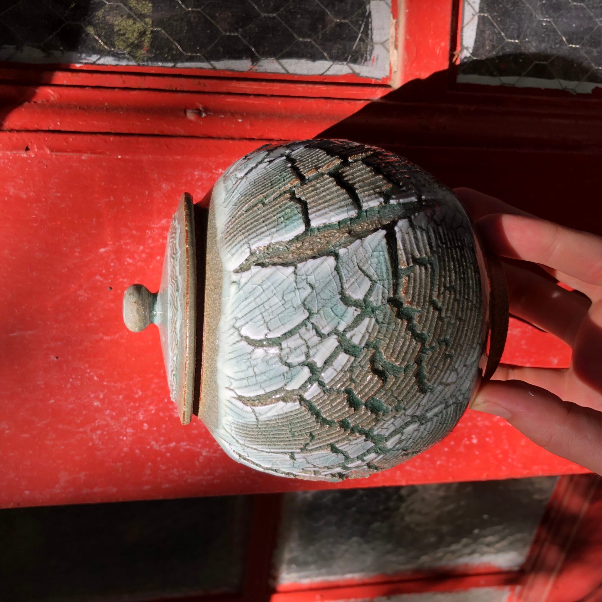 Person holding a cracked, weathered ceramic jar with a lid, against a red background, with visible cracks and peeling paint.