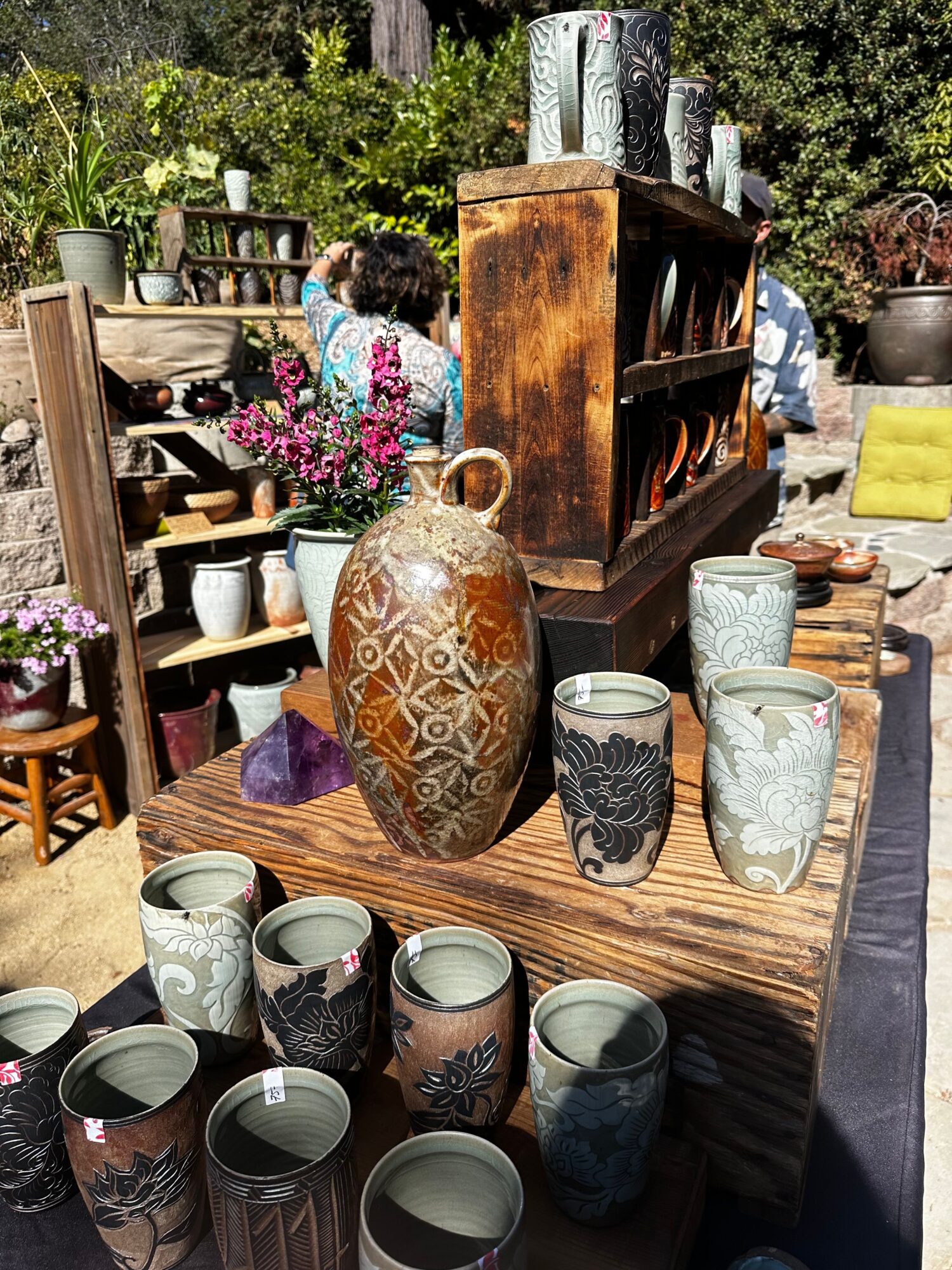Wooden table with ceramic vases and cups, surrounded by furniture and plants outdoors, with a person in the background.