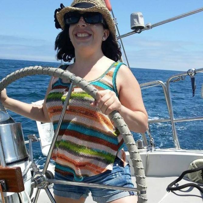 Woman smiling on a boat, wearing sunglasses, a straw hat, and a colorful striped top, holding the steering wheel.
