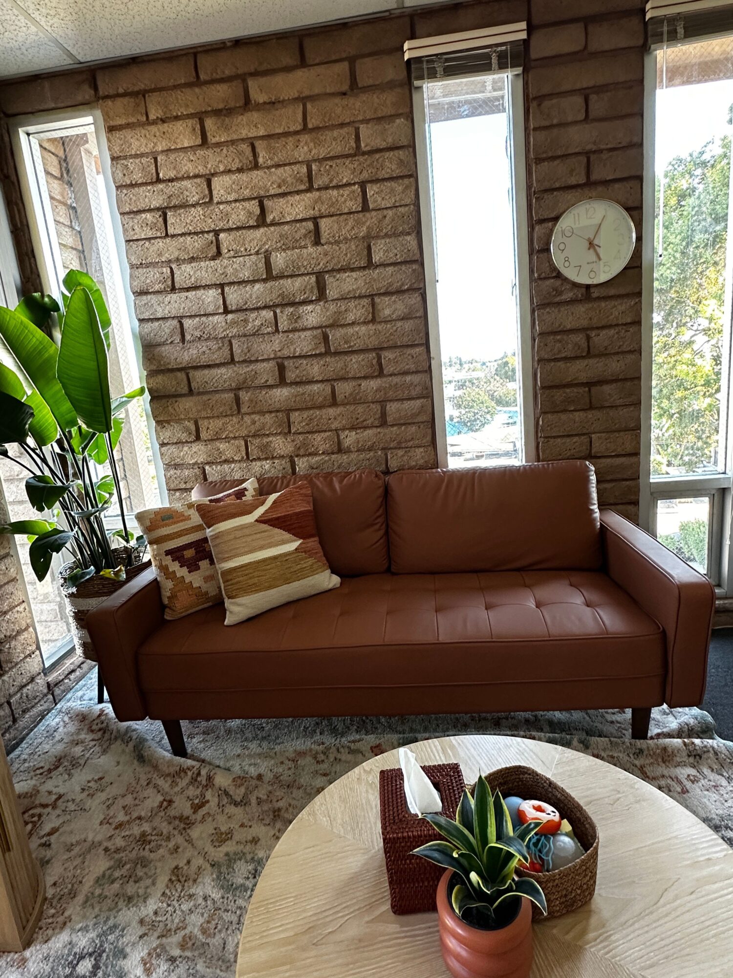 Living room with brick wall, window, clock, brown sofa, round wooden table with plant, and patterned rug.