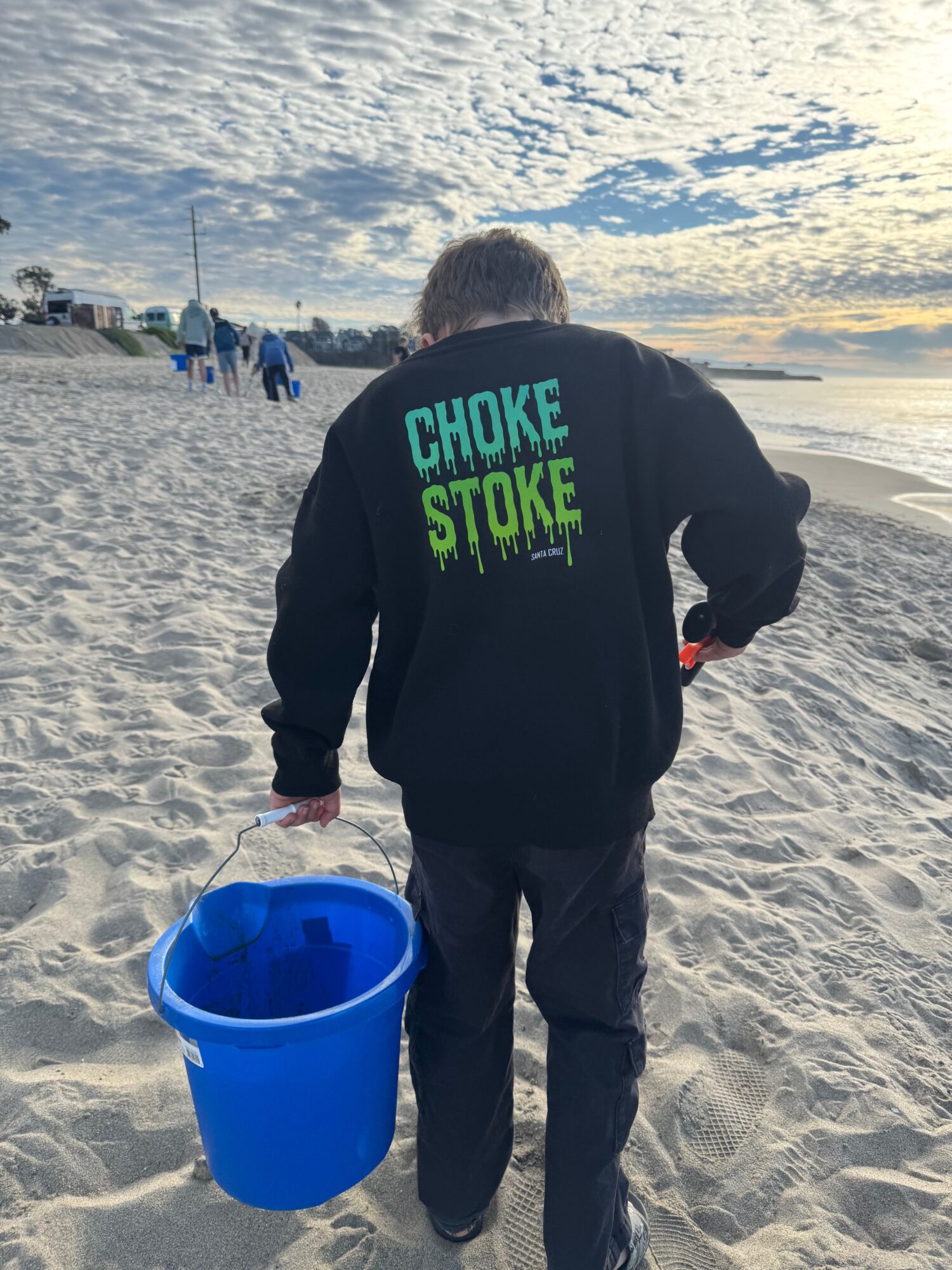 Person walking on sandy beach carrying a blue bucket, with clouds and ocean in the background.