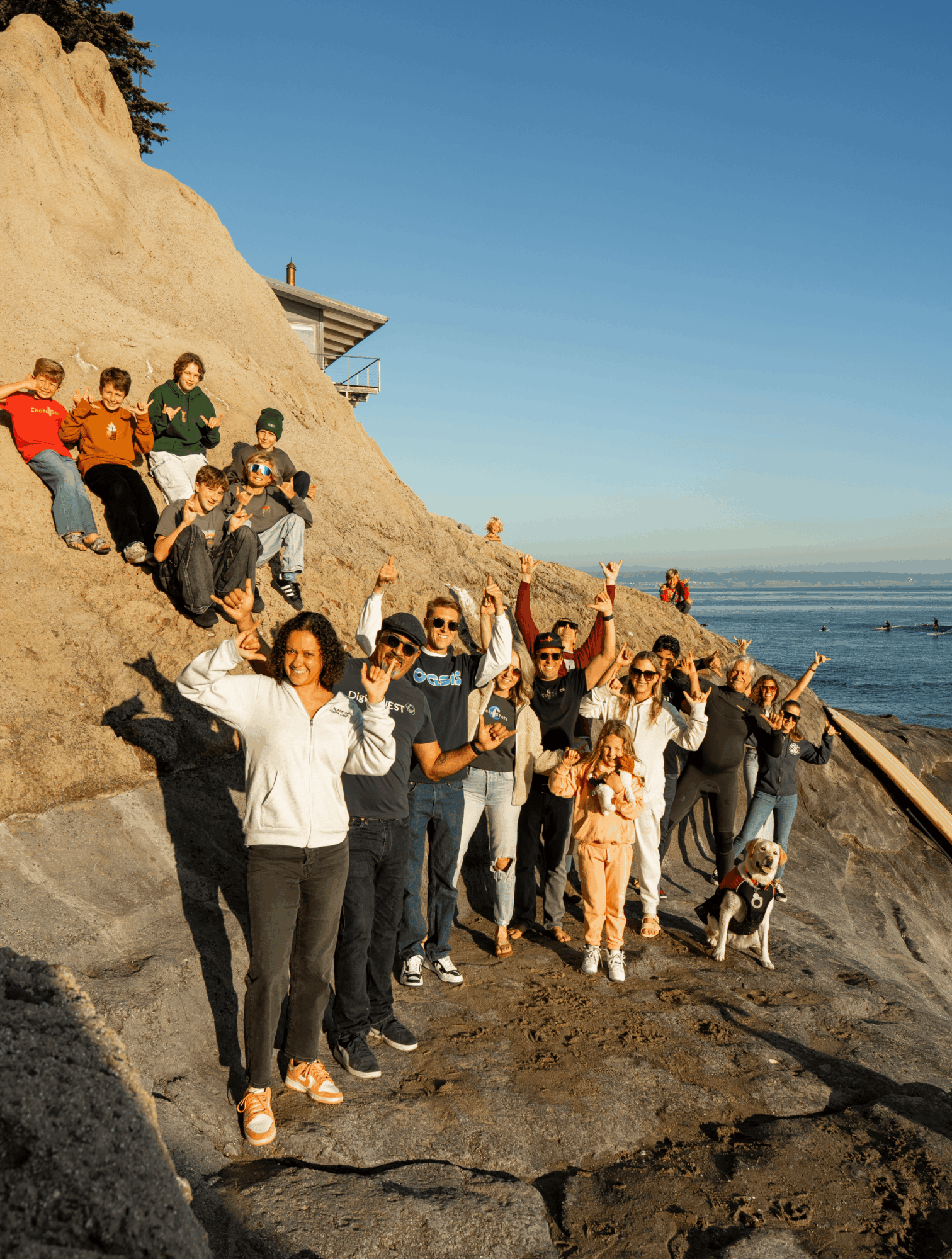 Group of people on a rocky beach hillside, some waving, with ocean and clear sky in background.