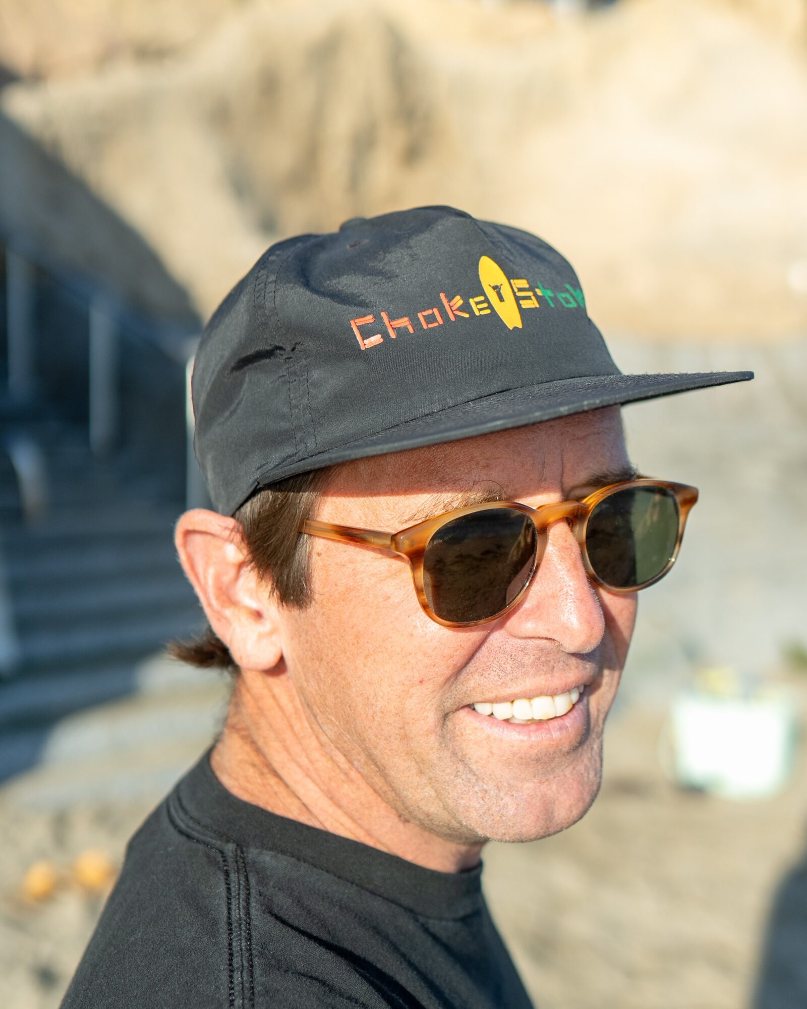 Man wearing sunglasses and a black cap with colorful logo outdoors, smiling.