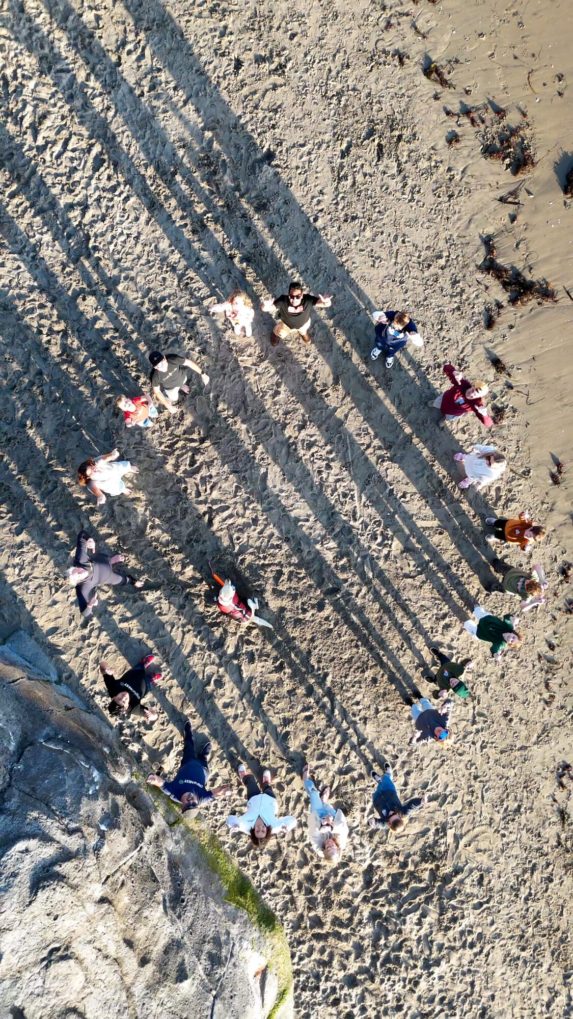People form a circle on sandy beach, casting long shadows, with ocean waves nearby.