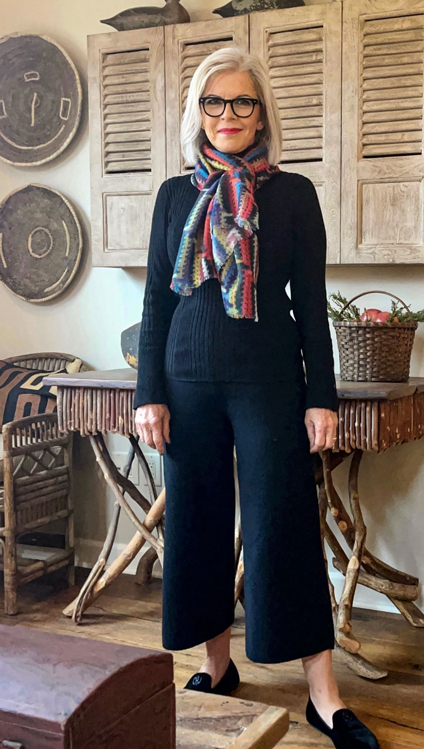 Woman with glasses and blonde hair wearing a dark sweater, colorful scarf, wide-leg pants, and black shoes, standing indoors.