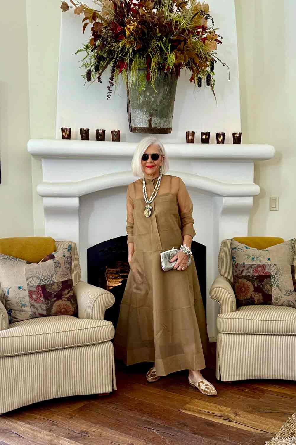 Older woman with white hair and sunglasses stands in front of a white fireplace with a large floral arrangement, flanked by two armchairs.