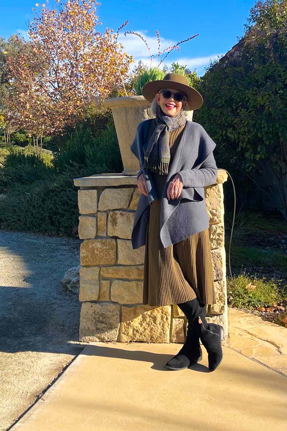 Woman in a wide-brimmed hat, sunglasses, gray coat, and brown skirt standing outdoors next to a stone pillar.