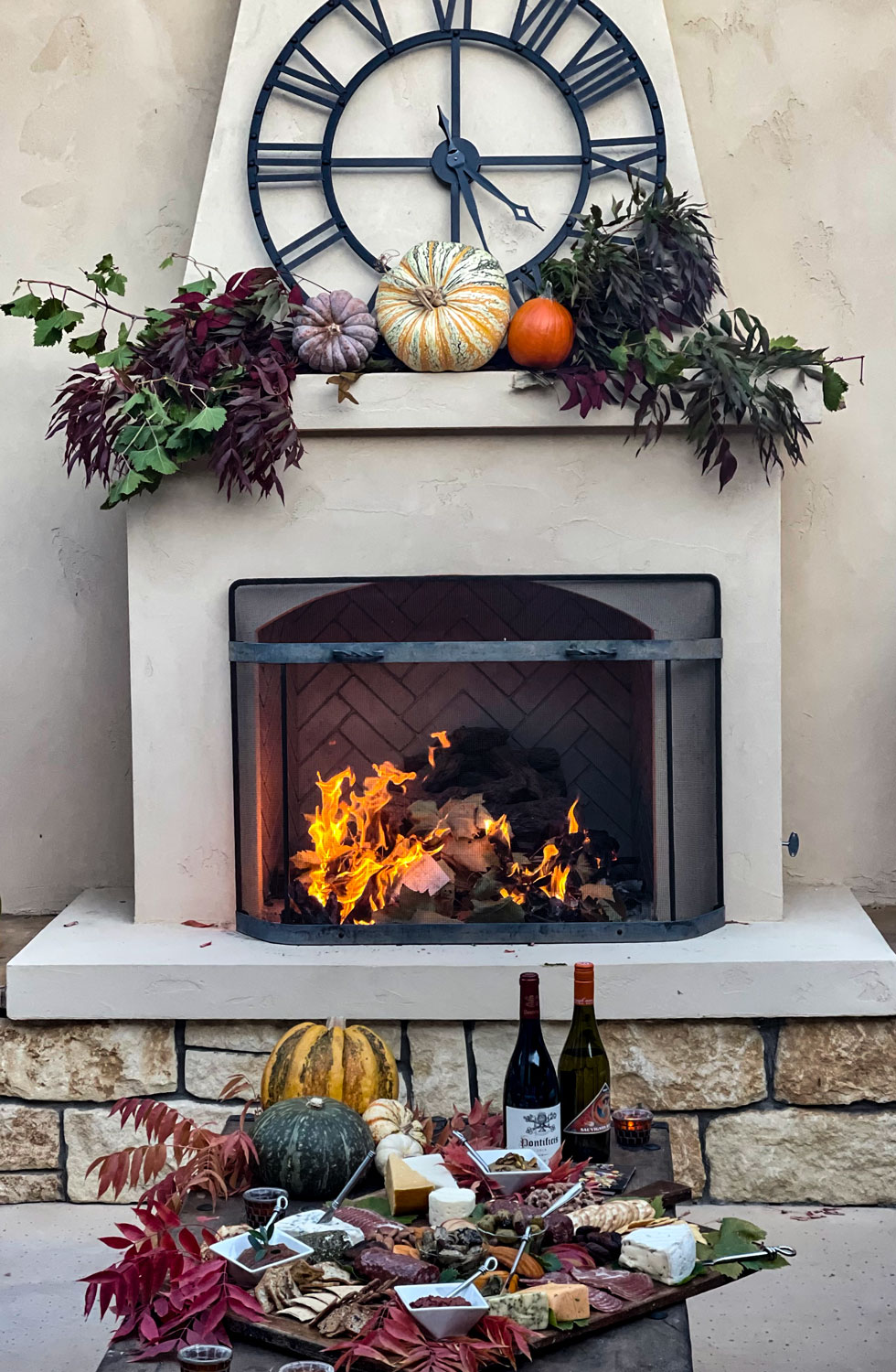 Fireplace with pumpkins, wine bottles, and autumn decorations, clock and floral arrangement above, stone and white mantel, cozy fall scene.