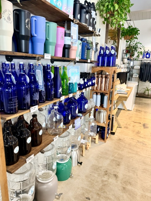 Shelves display colorful and blue bottles, with plants and furniture in the background, in a store setting.