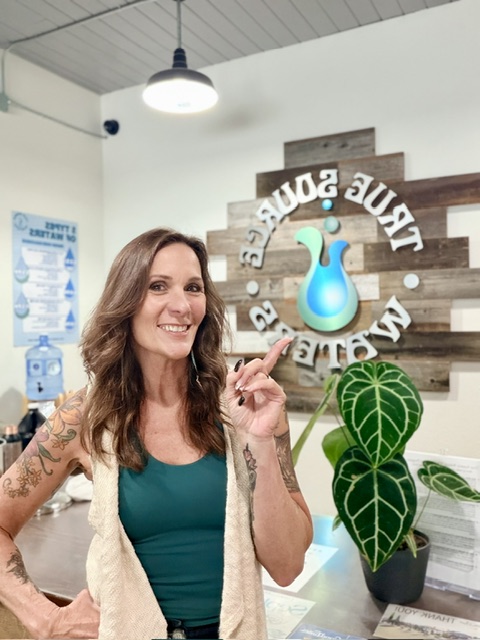 Woman with long brown hair smiling, pointing at a sign with a blue water droplet, in a room with a plant and wall decor.