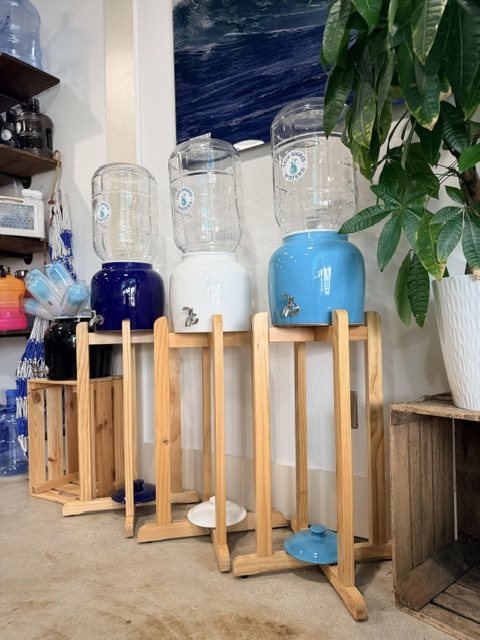 Four water dispensers on wooden stands, with a plant in a white pot nearby, inside a room with a window and shelves.