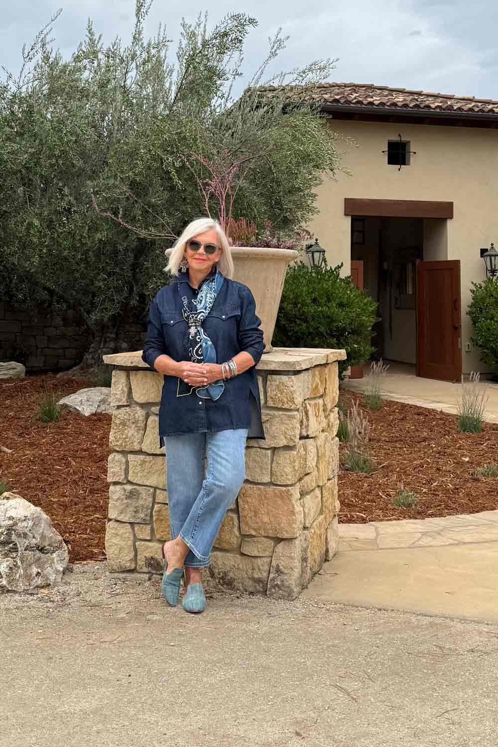 Woman with white hair and sunglasses leaning against a stone pillar outside a building, wearing a denim jacket and jeans.