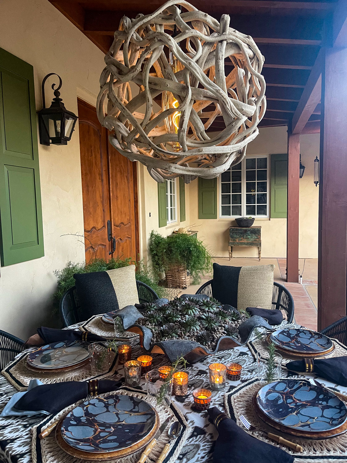 Outdoor dining table set with plates, glasses, and candles, under a large decorative hanging light fixture, with a patio background.