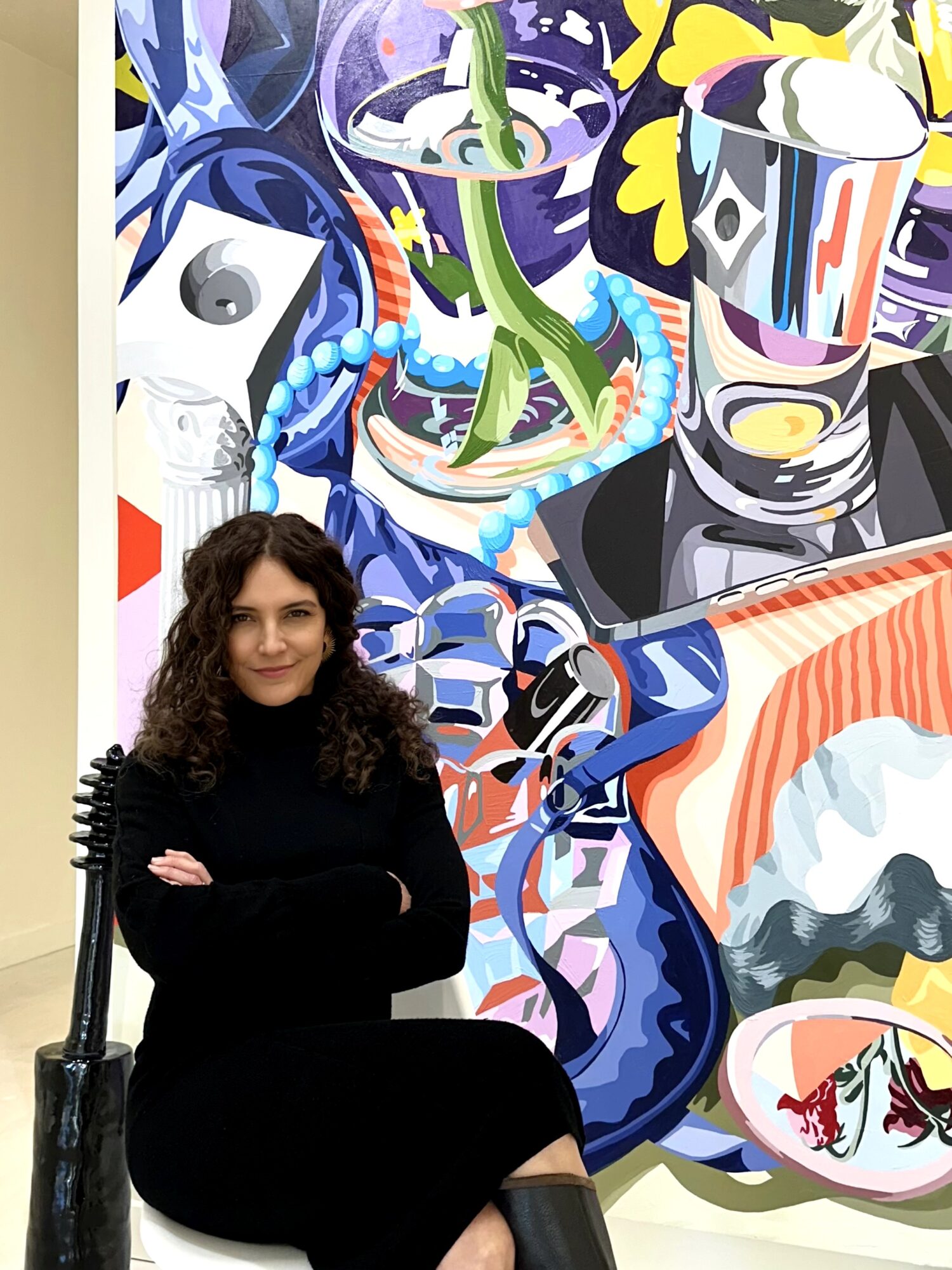Woman with curly hair in black clothing standing in front of a colorful abstract mural.