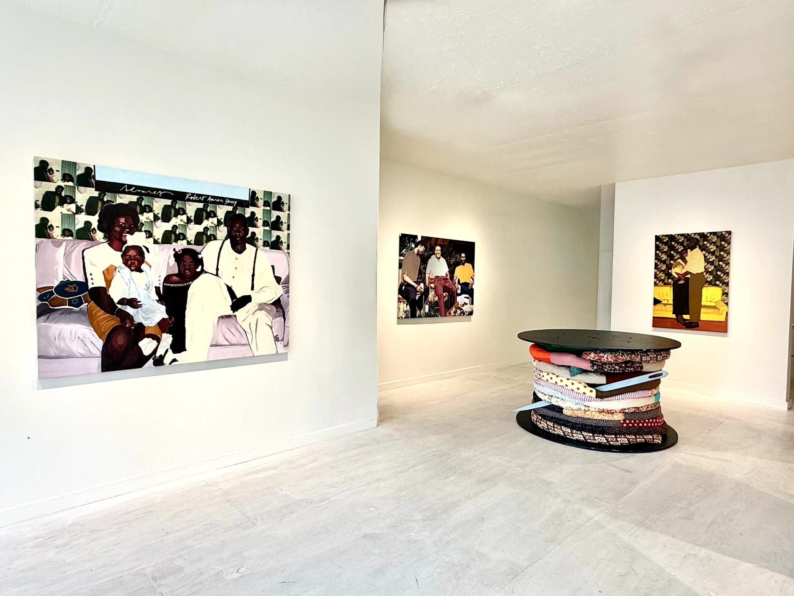 Art gallery with three paintings on white walls and a circular sculpture with stacked objects in the center.