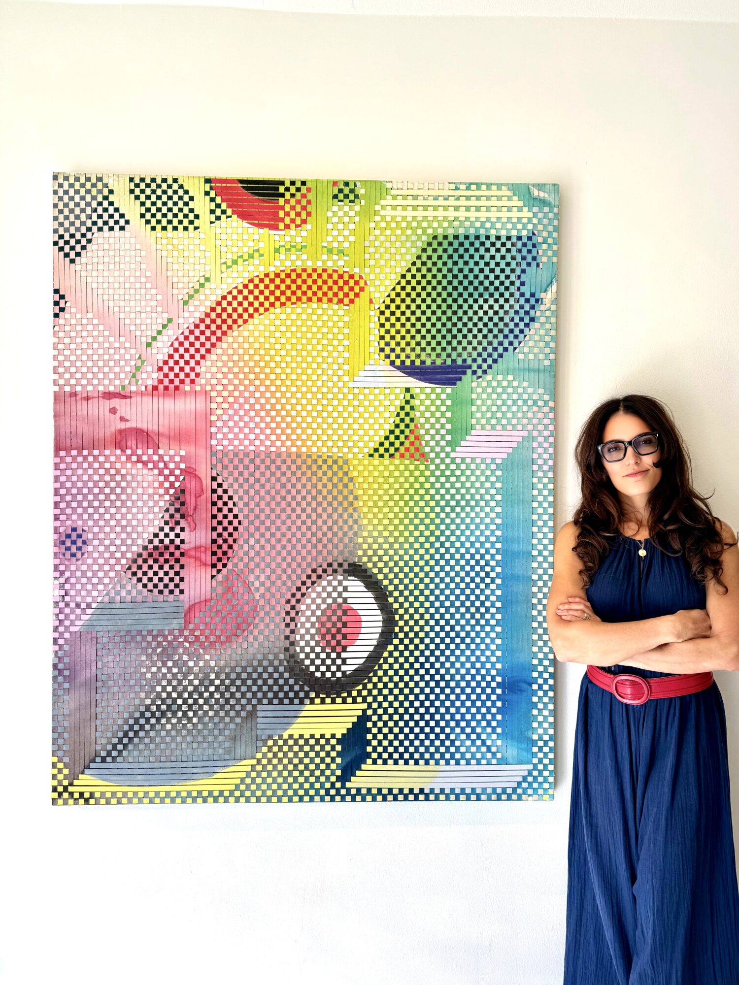 Woman with glasses and long hair standing with arms crossed, wearing a blue dress and red belt, against colorful artwork.