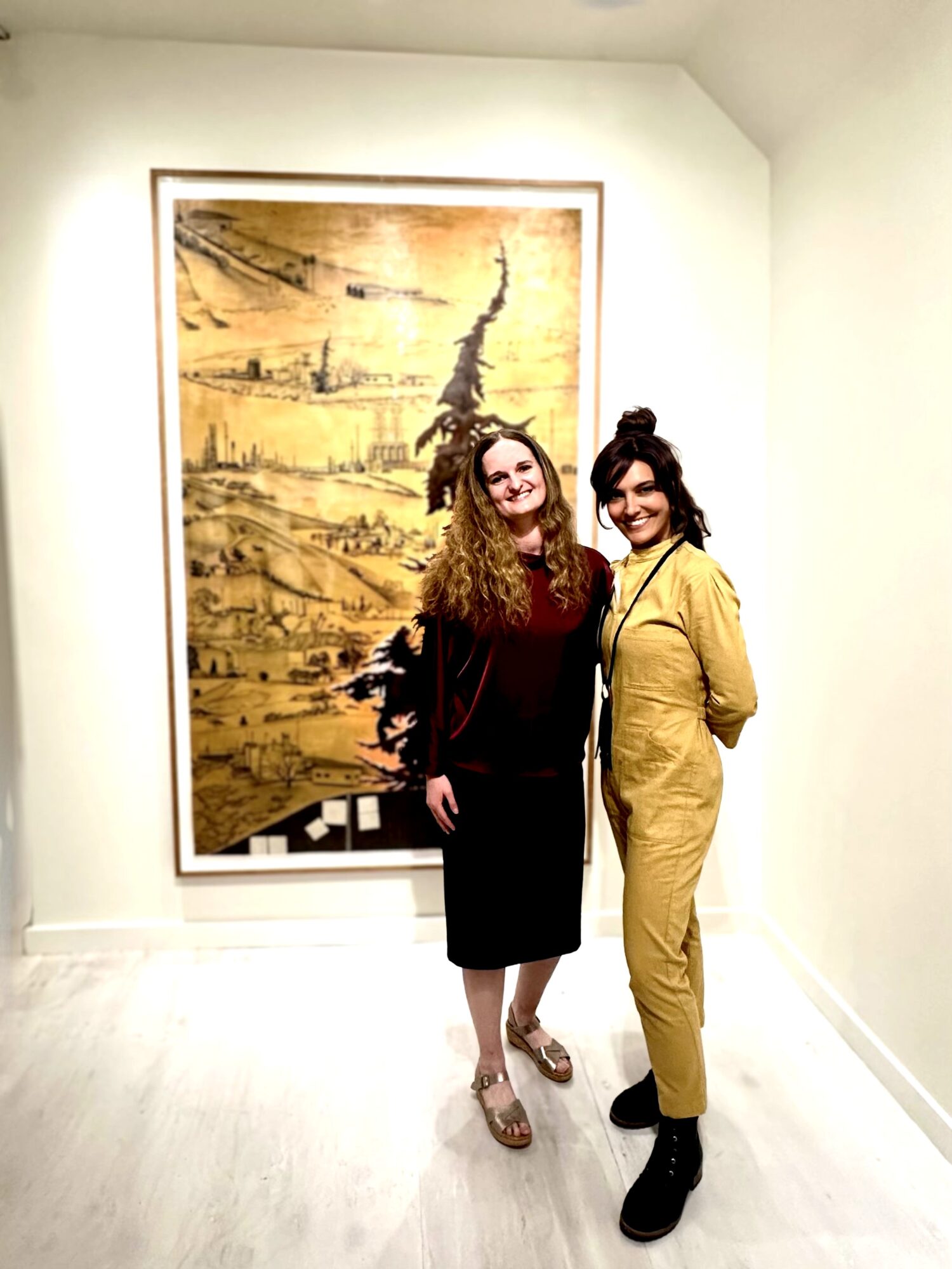 Two women stand in front of a large framed artwork on a white wall, smiling at the camera.