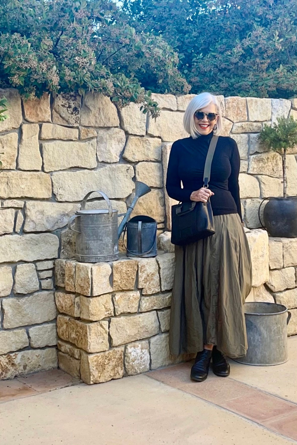 Woman with white hair and sunglasses standing outdoors against a stone wall, wearing a black top, long skirt, and black shoes.
