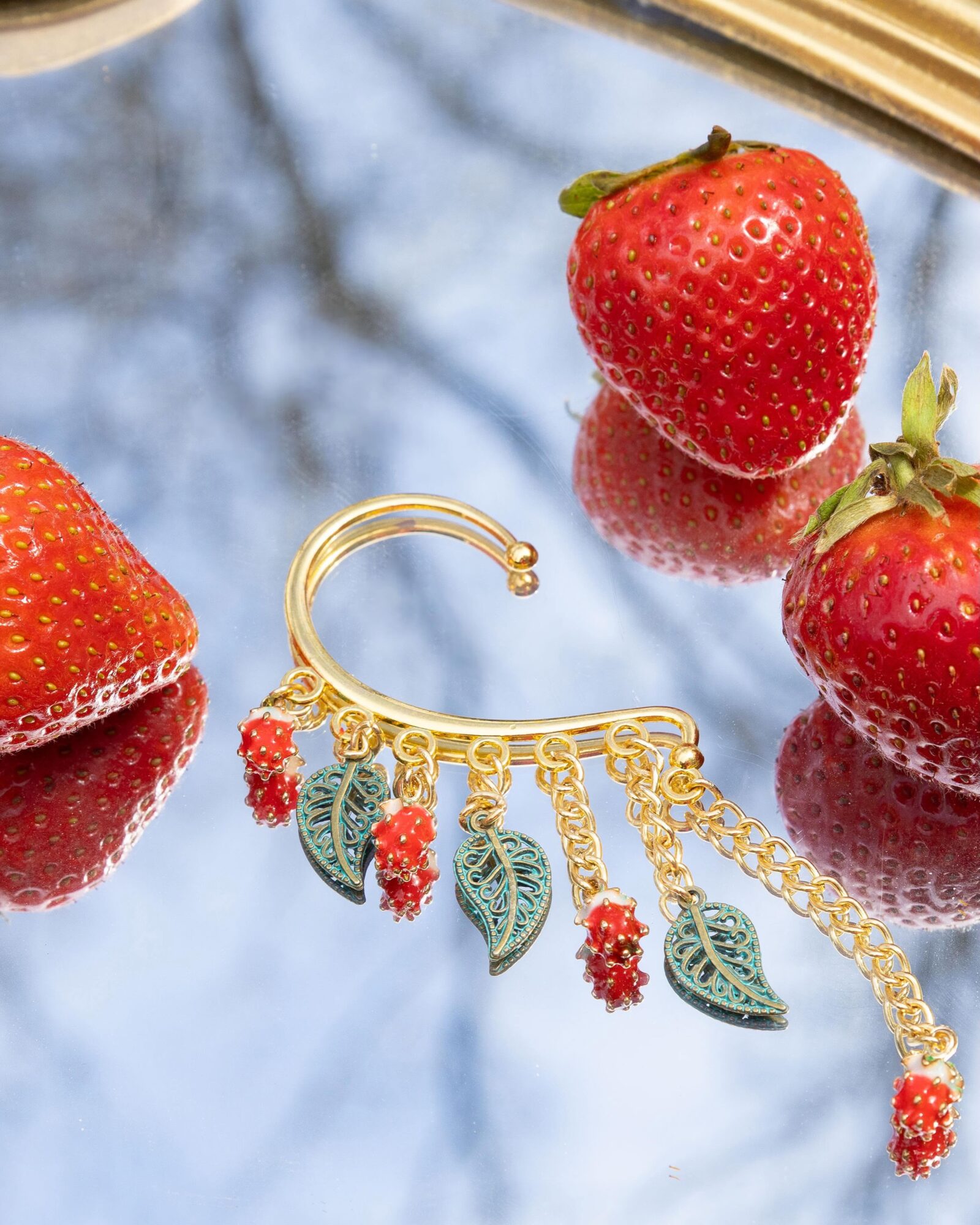 Three strawberries and a gold earring with leaf and red bead charms on a reflective surface.