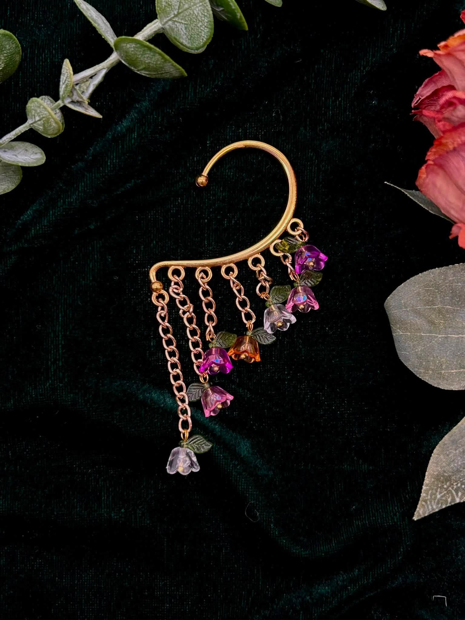 Gold earring with dangling purple and pink beads, hanging on dark fabric with green and pink leaves nearby.