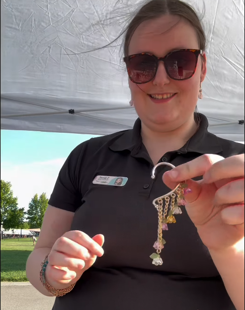 Woman outdoors holding a keychain with jewelry, wearing sunglasses and a black polo shirt, smiling.