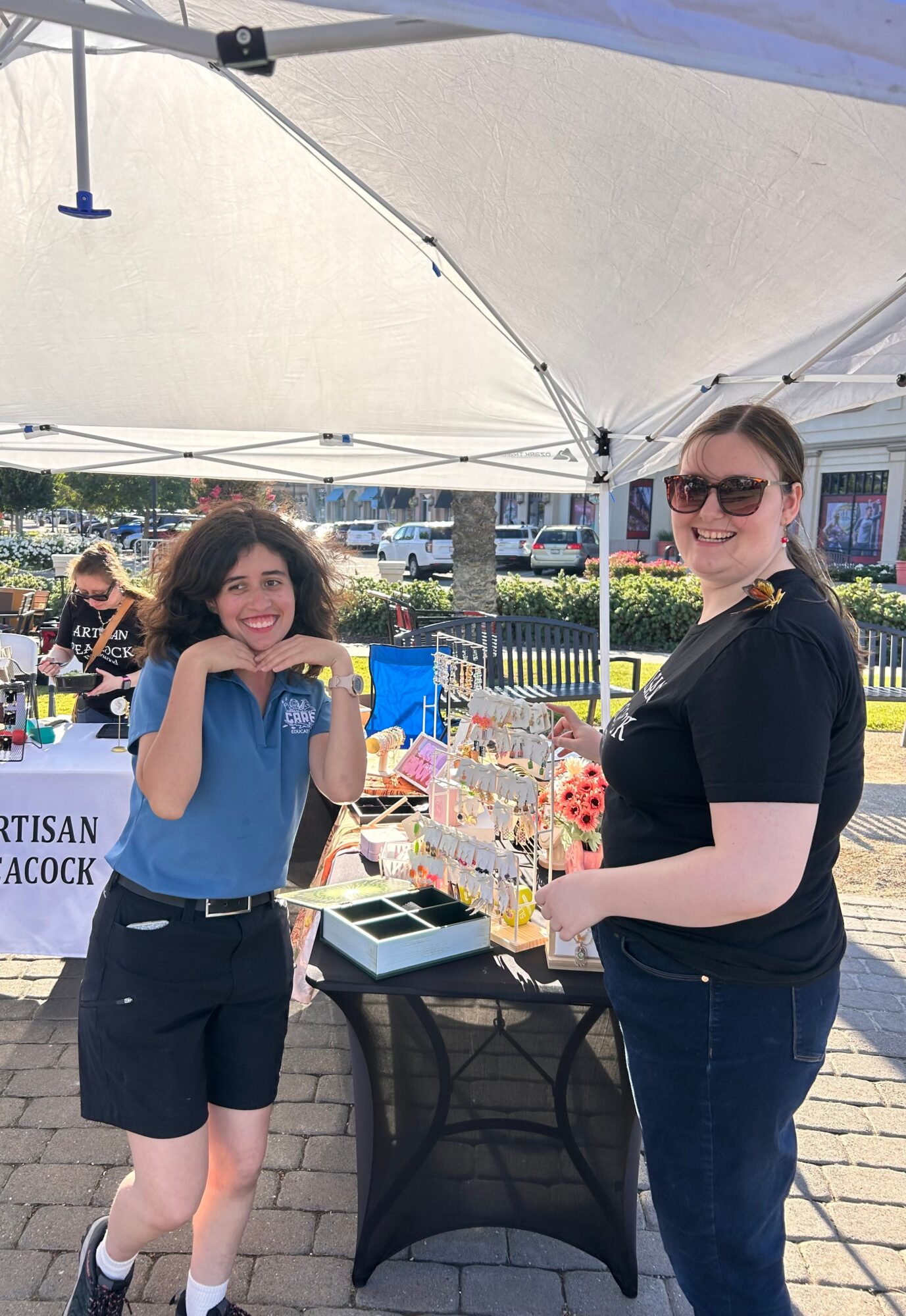 Two women standing outdoors under a canopy, smiling, with a table displaying colorful items, in a sunny setting.
