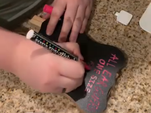 Person writes on a small black object with pink and white text, on a speckled countertop.