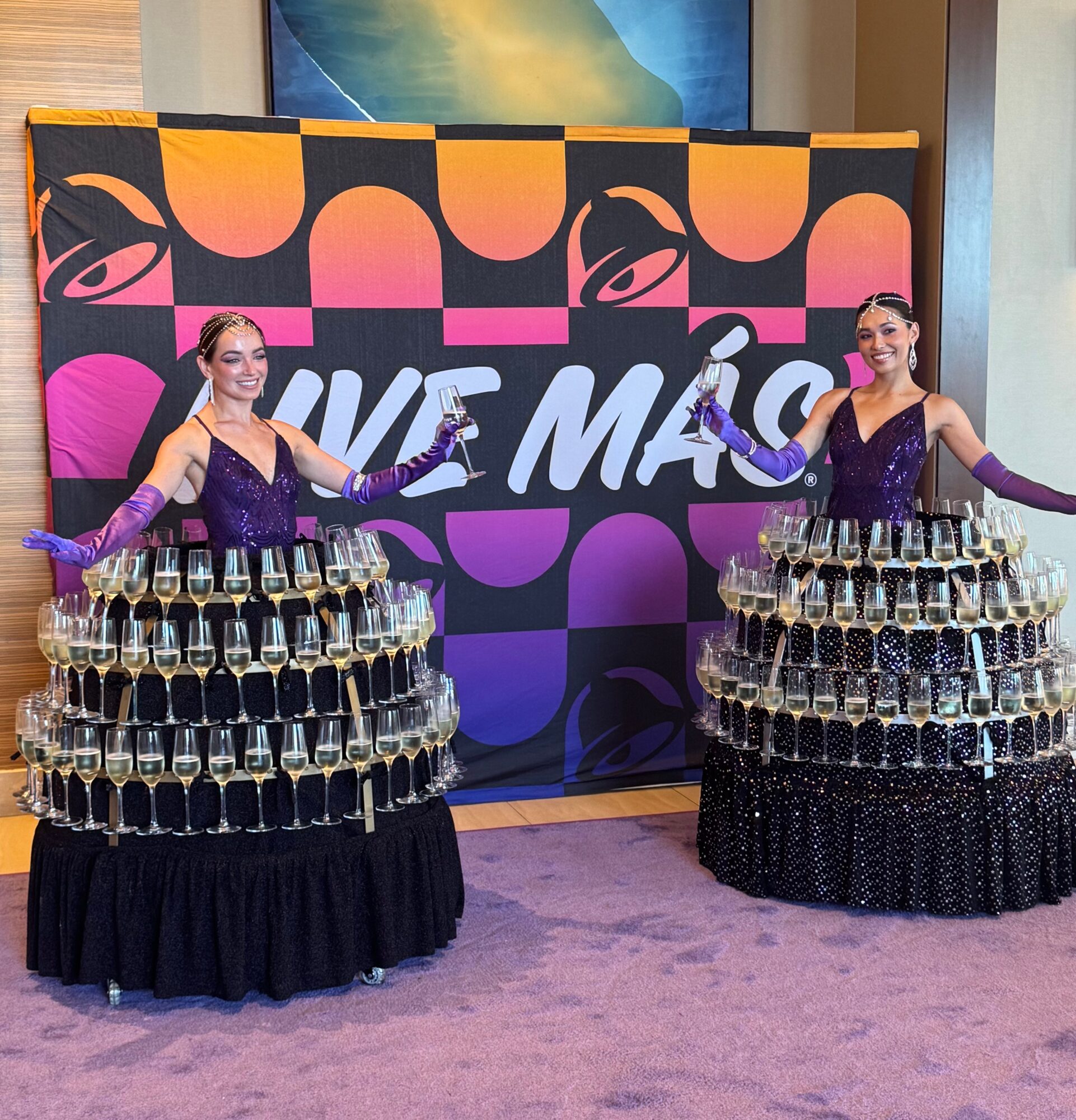 Two women in purple dresses stand behind tables with stacked glasses, smiling, in front of a colorful backdrop with text.